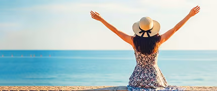 Woman in a straw sun hat and floral dress, seen from behind with arms outstretched, facing a calm blue sea under a bright sunlit sky