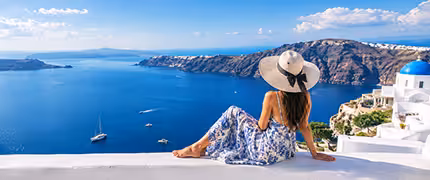 Woman in a sun hat and blue patterned dress sitting on a whitewashed wall in Santorini, looking out over the caldera with sailing boats on the deep blue sea