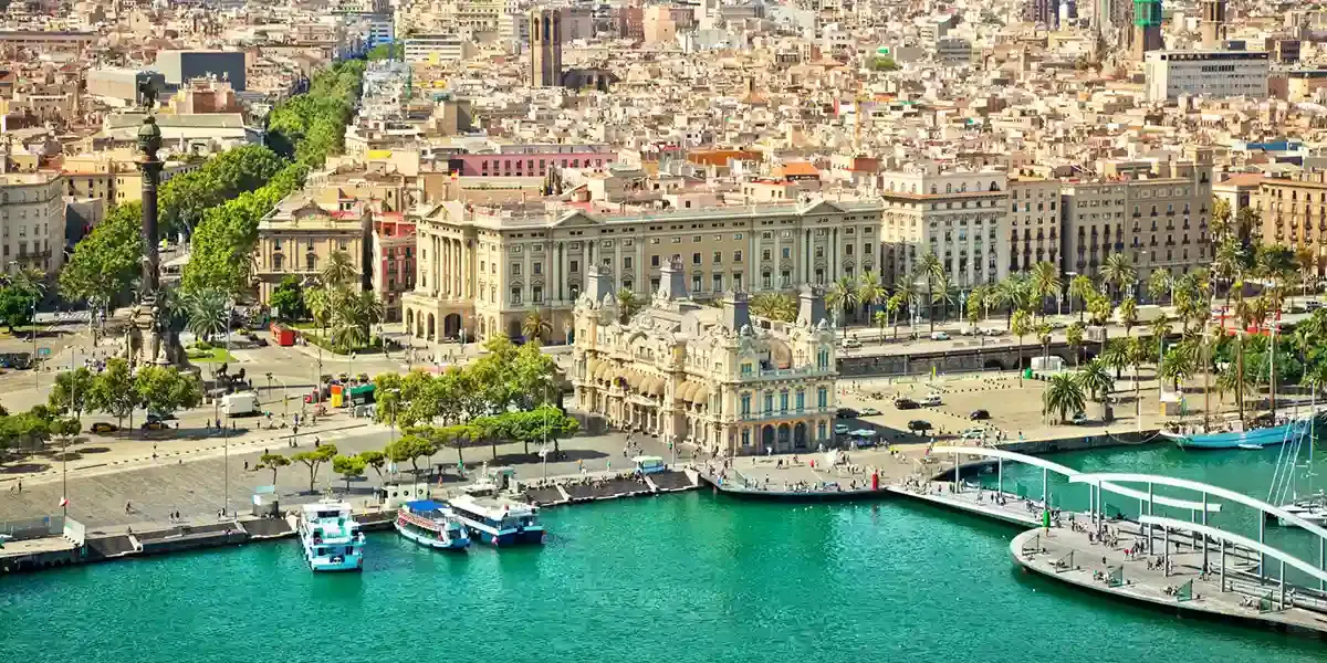 Aerial view of Barcelona's Port Vell waterfront, with the Columbus Monument, grand historic buildings, and turquoise harbour water