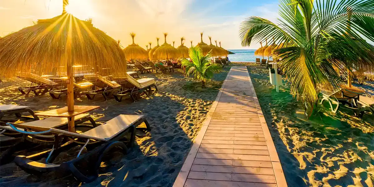 Sunrise over a tropical beach resort with thatched parasols, sun loungers, and a palm-lined wooden boardwalk