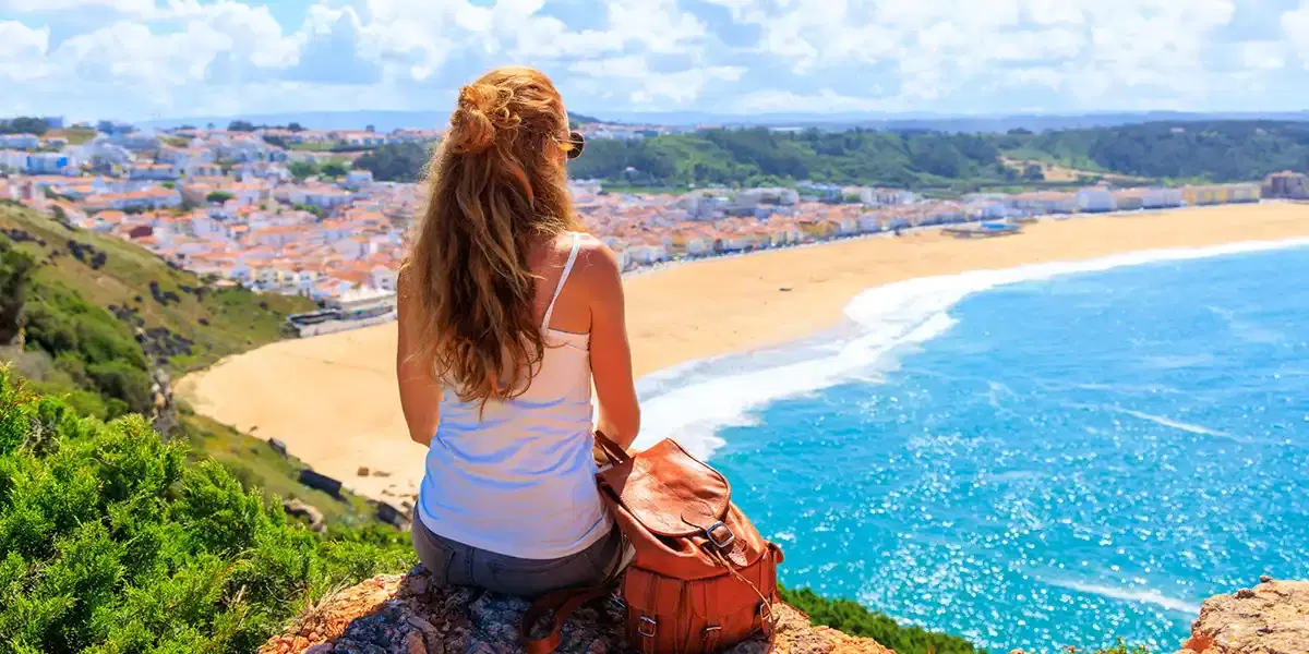 Woman with a backpack sitting on a clifftop overlooking a wide sandy bay and a whitewashed coastal town in Portugal