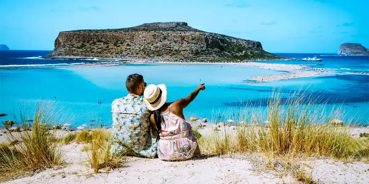 Couple sitting on a sand dune overlooking the turquoise Balos Lagoon with a rocky island in the distance