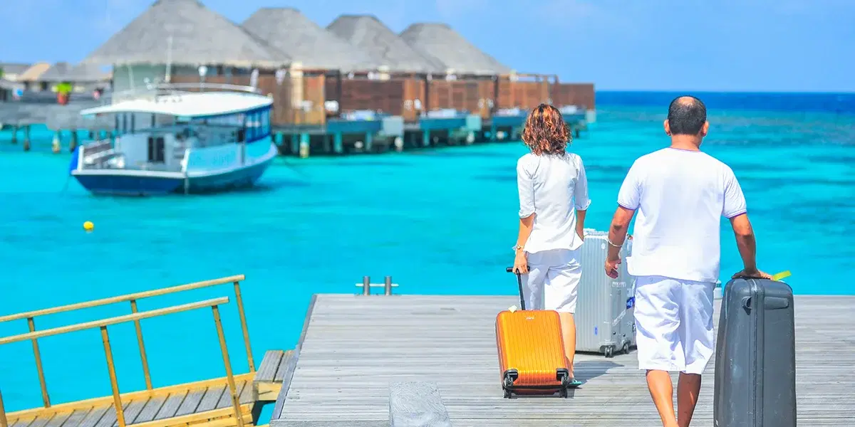 Couple pulling suitcases along a wooden jetty towards overwater bungalows above turquoise lagoon water